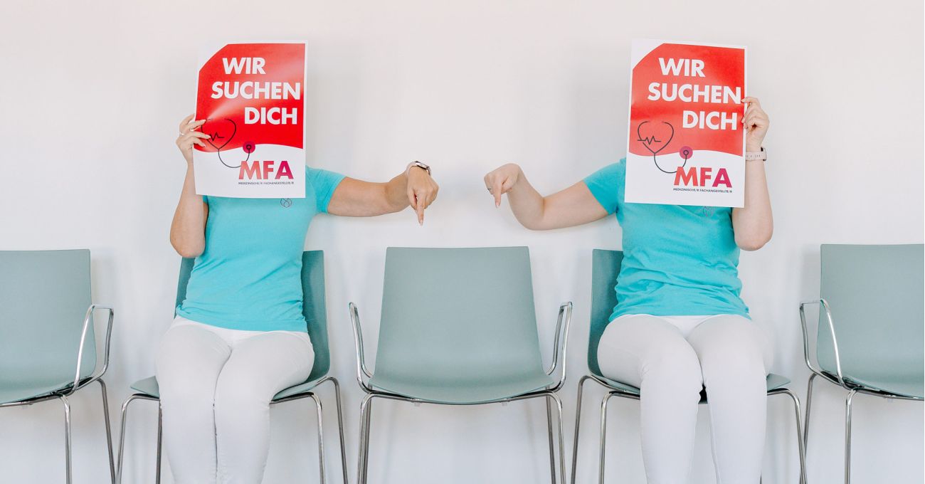 Zwei Mitarbeiterinnen der Herzpraxis Magdeburg halten Schilder „Wir suchen dich – MFA“ und zeigen auf einen freien Stuhl.