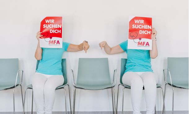Zwei Mitarbeiterinnen der Herzpraxis Magdeburg halten Schilder „Wir suchen dich – MFA“ und zeigen auf einen freien Stuhl.